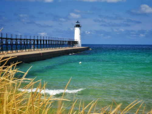 A white lighthouse stands on a pier by the turquoise sea, with gentle waves and grassy foreground under a blue sky.