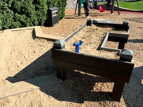 A wooden sandbox with a blue toy shovel, surrounded by sand and playground equipment in the background.
