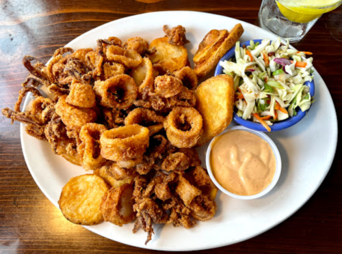 A plate of fried seafood, including calamari and onion rings, served with coleslaw and a dipping sauce.