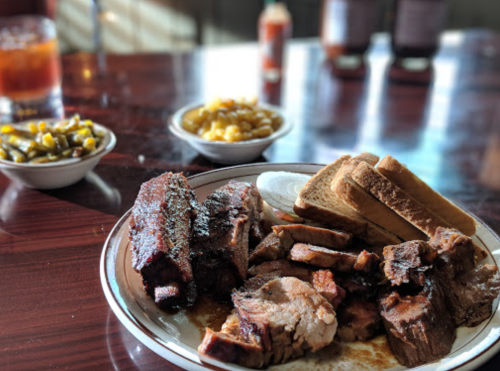 A plate of barbecue ribs and sliced meat, served with bread and sides of green beans and corn.
