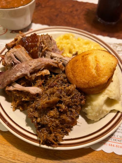 A plate of pulled pork, cornbread, macaroni and cheese, and mashed potatoes, with a side of beans in the background.