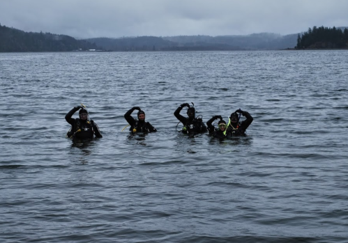 Four divers in wetsuits are standing in a lake, waving and smiling at the camera. Overcast skies in the background.