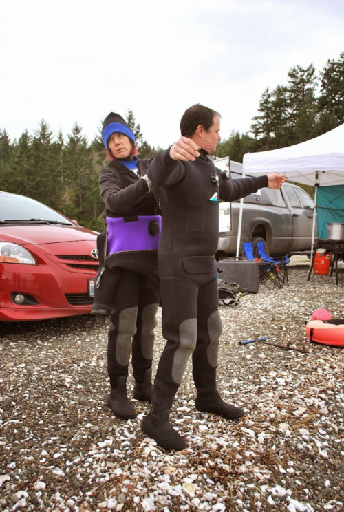 Two people in wetsuits, one assisting the other with their suit, set against a backdrop of trees and parked vehicles.