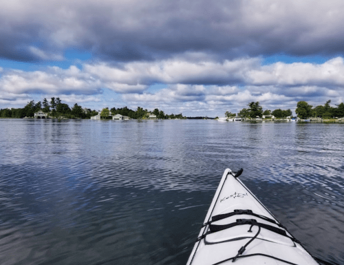 A kayak glides over calm water, surrounded by lush greenery and cloudy skies.