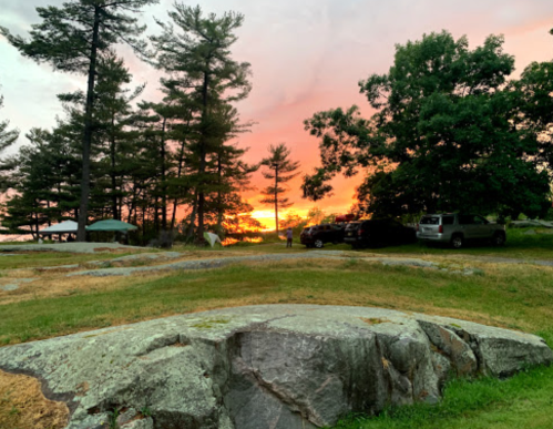 A serene landscape featuring trees and rocks, with a vibrant sunset in the background and parked cars nearby.