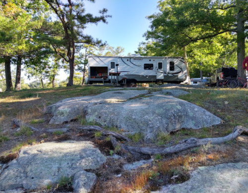 A camper parked among trees and large rocks, with a clear blue sky in the background.