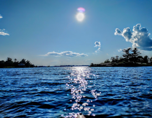 A serene view of a sunlit lake with sparkling water and a clear blue sky dotted with clouds.