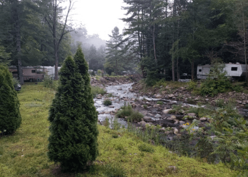 A serene campsite by a rocky stream, surrounded by trees and RVs, with mist in the background.