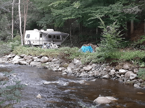A camper parked by a rocky riverbank, surrounded by trees and greenery.
