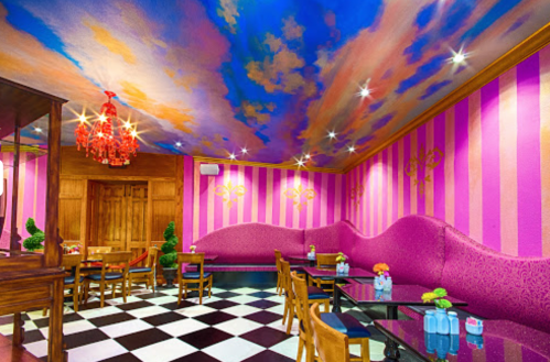 Colorful café interior with a painted ceiling, pink striped walls, a plush curved seating area, and checkered flooring.