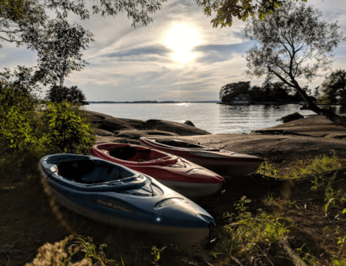 Three kayaks in blue, red, and gray rest on a rocky shore, with a serene lake and sunset in the background.
