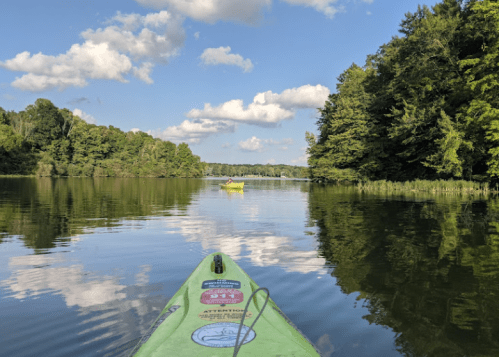 A kayak on calm water, surrounded by lush green trees and a blue sky with fluffy clouds.