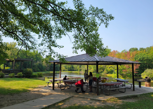 A shaded picnic area by a lake, with people sitting at tables and greenery in the background.