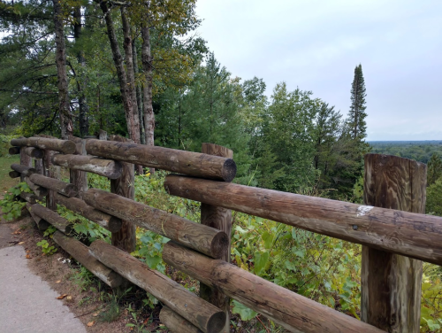 A wooden fence along a path, surrounded by lush green trees and a cloudy sky in the background.