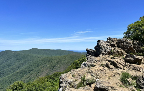 A rocky outcrop overlooks a lush green mountain range under a clear blue sky.