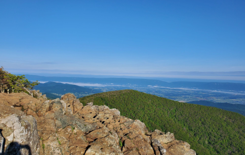 A panoramic view from a rocky mountain peak, showcasing lush green hills and a clear blue sky.