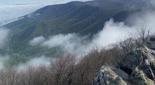 A rocky outcrop overlooks lush green mountains shrouded in mist and clouds under a clear blue sky.