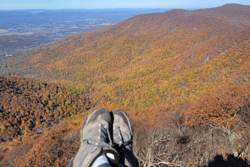 A pair of hiking boots resting on a rocky ledge, overlooking a vibrant autumn landscape of mountains and valleys.