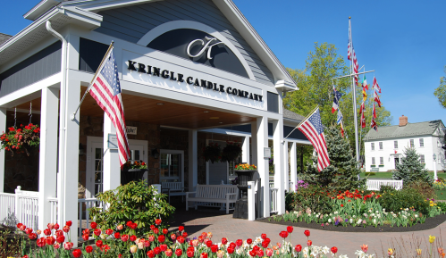 Exterior of Kringle Candle Company with American flags, colorful flowers, and a clear blue sky.