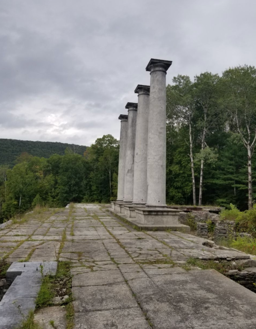 Four tall, weathered columns stand on a stone platform, surrounded by trees and a cloudy sky.