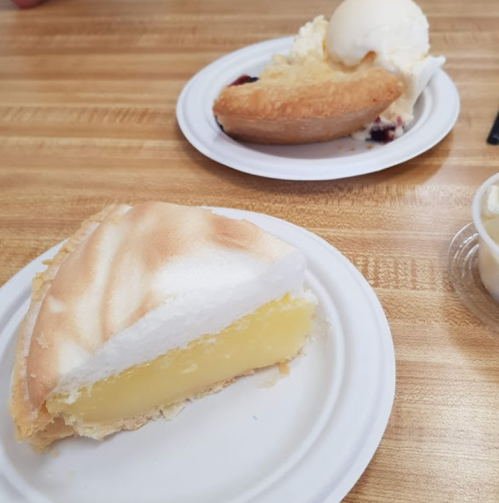 A slice of lemon meringue pie on a paper plate, with another dessert and ice cream in the background.