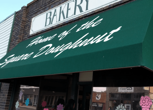 A bakery storefront with a green awning that reads "Home of the Square Doughnut." Colorful decorations in the windows.