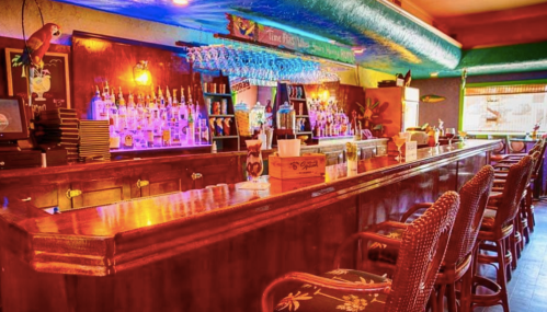 A vibrant bar with colorful bottles, wooden counter, and rattan chairs, featuring tropical decor and a parrot.