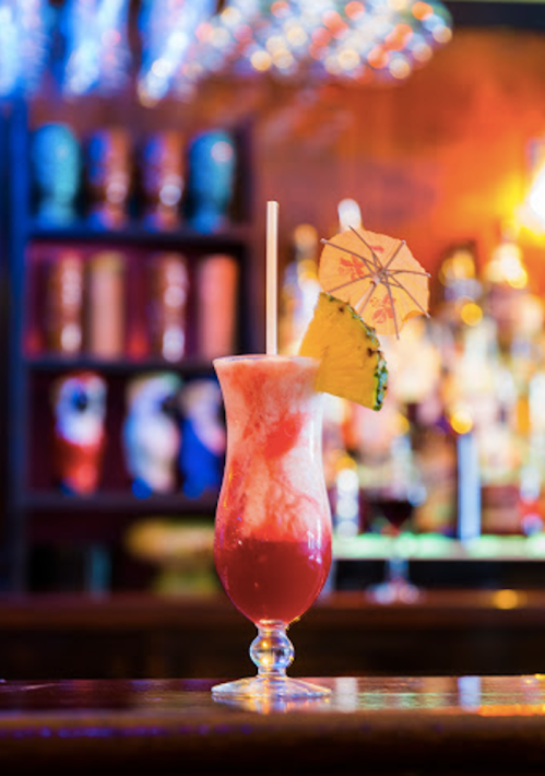 A colorful cocktail with a pineapple slice and an umbrella, set against a blurred bar background.