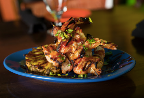 A vibrant blue plate with grilled shrimp topped with sauce, served on a bed of grilled pineapple slices.