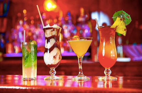 Four colorful cocktails on a bar, including a dessert drink, a martini, and tropical beverages, with a vibrant background.