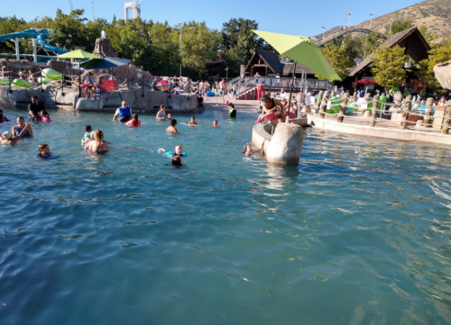 A busy water park scene with people swimming and lounging by the pool under green umbrellas.