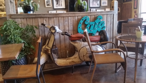 A vintage scooter sits beside a wooden counter in a cozy café, surrounded by chairs and plants.