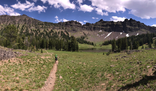 A panoramic view of a lush green valley with a winding trail, surrounded by mountains and a serene lake under a blue sky.