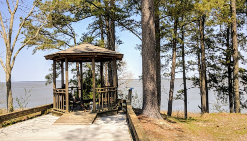 A wooden gazebo surrounded by trees, overlooking a calm body of water on a sunny day.