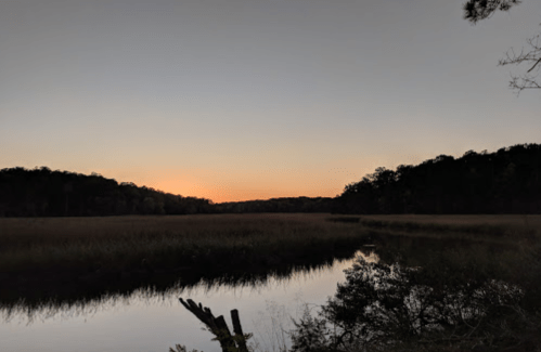 A serene landscape at dusk, featuring a calm river, grassy marsh, and silhouetted trees against a fading orange sky.