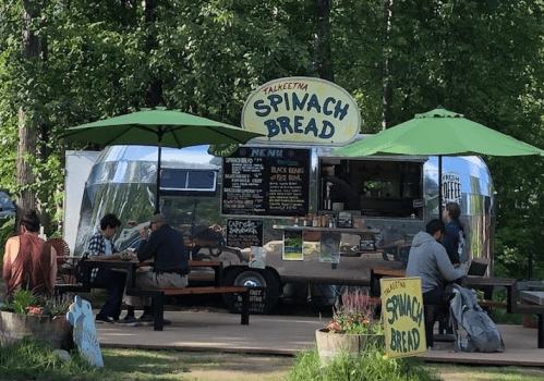 A food truck named "Talketna Spinach Bread" with green umbrellas, serving customers seated at outdoor tables.