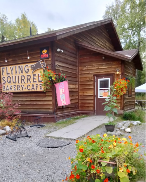 A rustic wooden café with a sign reading "Flying Squirrel Bakery-Café" and colorful flowers in front.