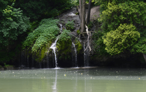 A serene scene of a small waterfall cascading over rocks into a calm, reflective body of water surrounded by lush greenery.