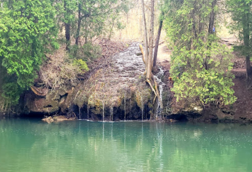 A serene scene of a small waterfall cascading over rocks into a calm, greenish-blue pond, surrounded by trees.