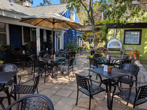 Outdoor patio with black metal tables and chairs, surrounded by greenery and colorful flower pots. Umbrella provides shade.