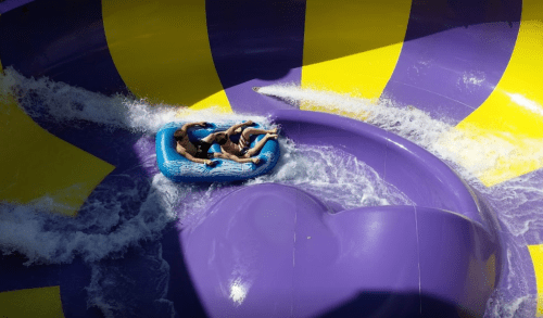 Two people ride a blue inflatable raft down a colorful water slide, surrounded by splashing water.