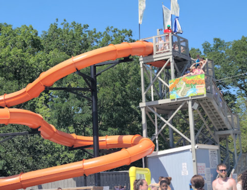 A bright orange water slide towers over a pool area, with people preparing to slide down on a sunny day.