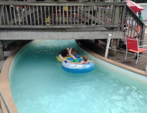 Two children float on colorful inner tubes in a lazy river under a wooden structure.