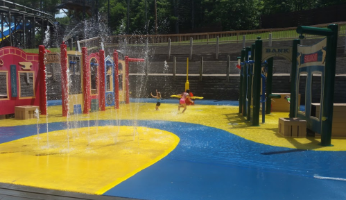 Children play in a colorful water park with fountains and bright yellow and blue surfaces.