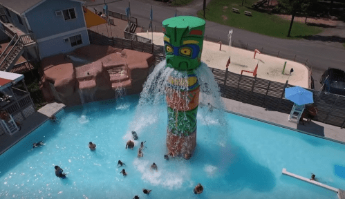 A colorful totem pole fountain sprays water into a pool filled with people enjoying a sunny day.