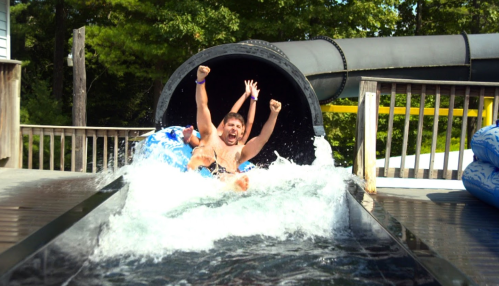 Two excited people slide down a water slide, splashing into the pool below with arms raised in joy.