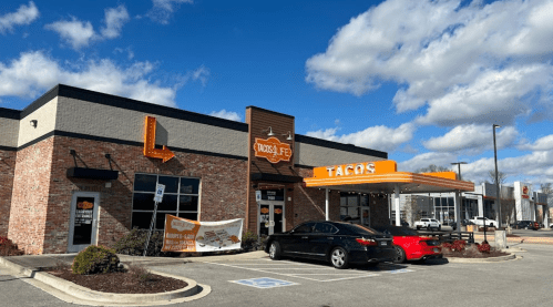 Exterior of a taco restaurant with a brick facade, orange awning, and parked cars under a partly cloudy sky.