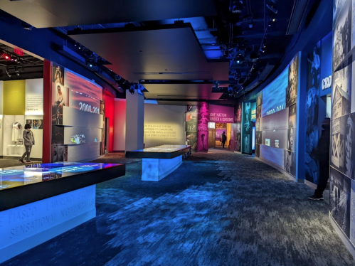 A modern exhibition space with colorful walls displaying text and images, featuring interactive displays and visitors exploring.