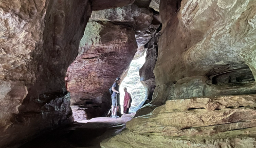 Two people stand inside a large, rocky cave, surrounded by colorful stone formations and natural light.