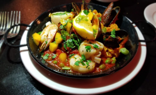 A vibrant seafood dish with shrimp, squid, mussels, and vegetables, garnished with lemon and herbs in a black skillet.